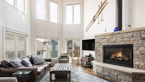 A spacious living room with a stone fireplace, leather sofa, and a large window overlooking a house.