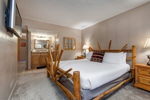 A bedroom with a wooden bed frame, a bedside table, a nightstand, a mirror, and a built-in TV.