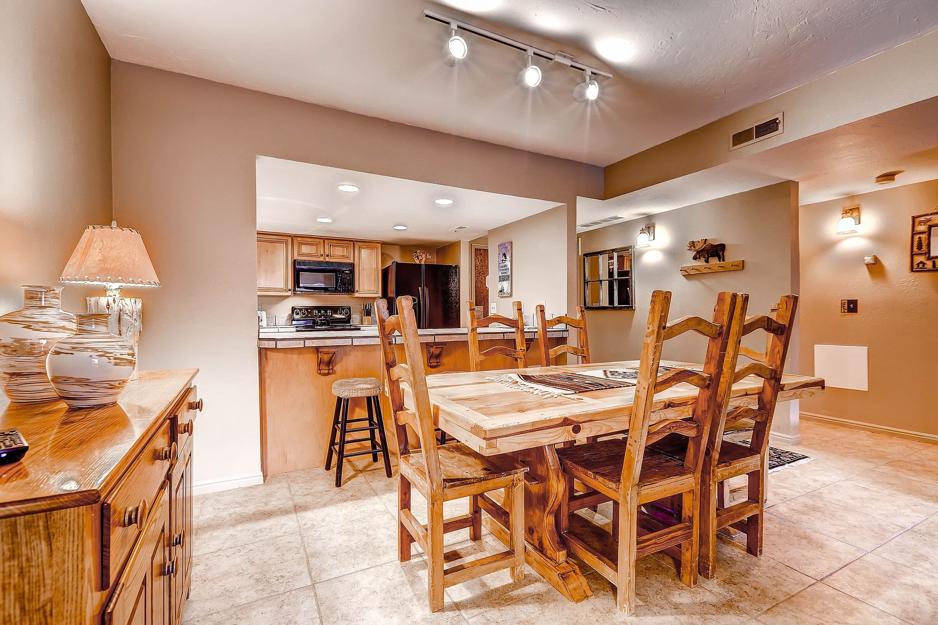A dining area with a wooden table and chairs, a lamp, and a kitchen in the background.