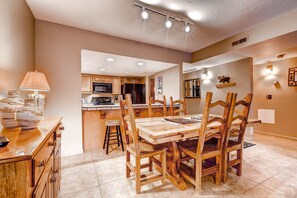 A dining area with a wooden table and chairs, a lamp, and a kitchen in the background.