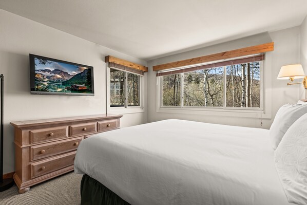 A bedroom with a bed, dresser, TV, and windows with a view of trees and mountains.