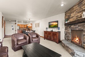 Una sala de estar con sillones de cuero, una mesa de centro, chimenea de piedra, televisor y una cocina al fondo.