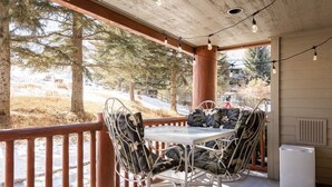 A covered outdoor seating area with a table and chairs, a refrigerator, and a view of a snowy landscape with trees.
