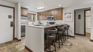 A modern kitchen with wooden cabinets, a stainless steel refrigerator, a microwave, and a bar area with stools. - Snow Flower 056 3 Bedroom Condo by RedAwning (Park City)