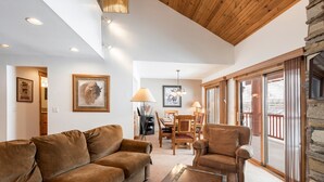 A cozy living room with a brown sofa, a wicker chair, and a dining table. There are framed pictures on the walls and a chandelier hanging from the ceiling. - Snow Flower 074 3 Bedroom Condo by RedAwning (Park City)