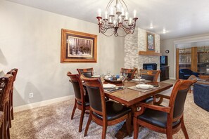 Uma sala de jantar com mesa de madeira, cadeiras e um lustre. Há uma lareira e uma janela grande ao fundo.