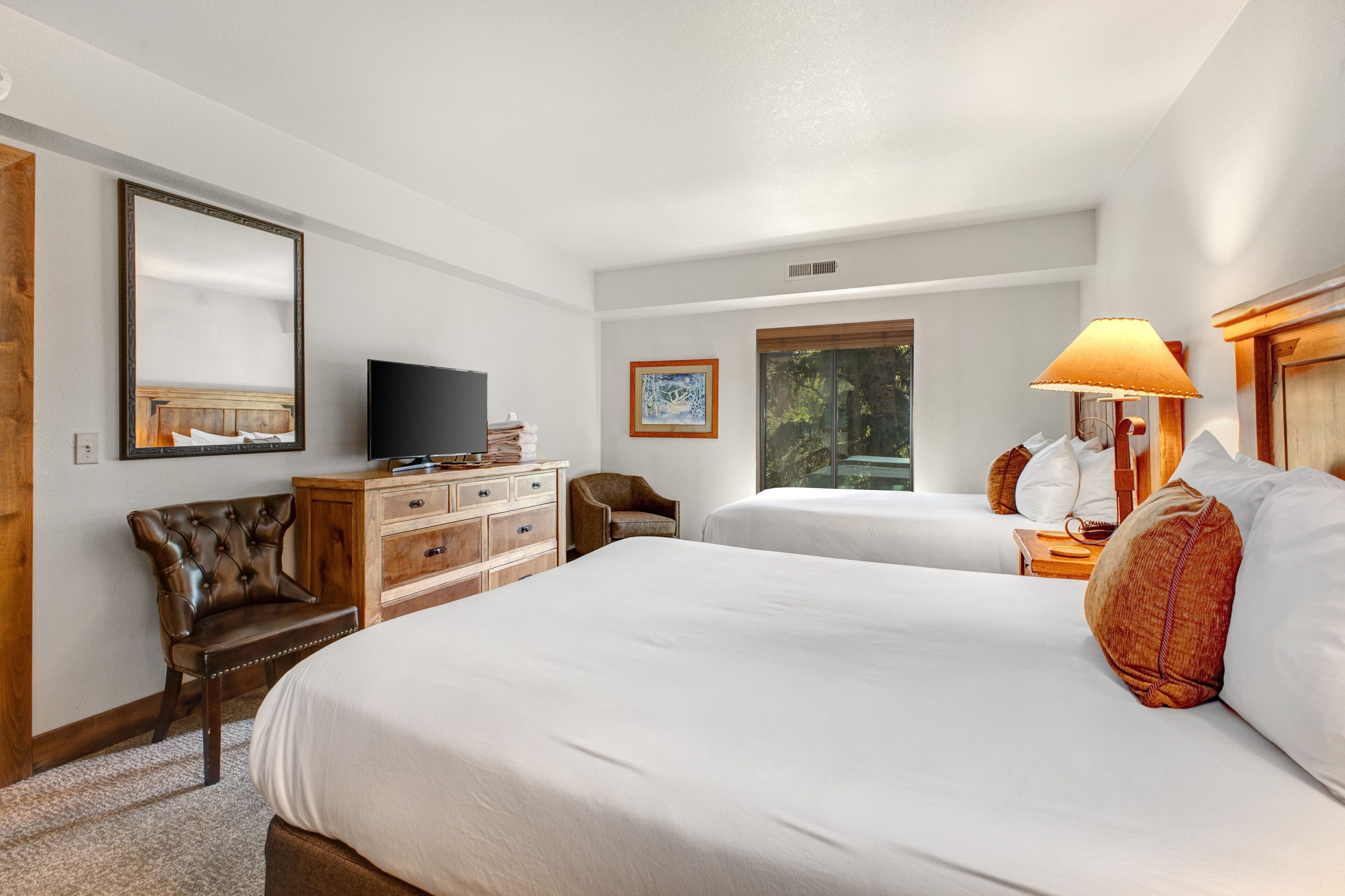 Ett hotellrum med två sängar, en tv, en byrå i trä, en stol och ett fönster med utsikt över träd.