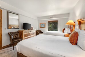Ein Hotelzimmer mit zwei Betten, einem Fernseher, einer Holzkommode, einem Stuhl und einem Fenster mit Blick auf Bäume.