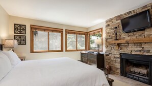 A bedroom with a large bed, a stone fireplace, a television mounted on the wall, and two windows with wooden blinds. - Snow Flower 023 2 Bedroom Condo by RedAwning (Park City)