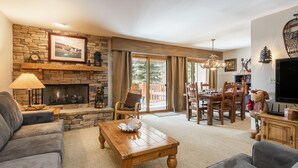 A cozy living room with a stone fireplace, a dining area with wooden furniture, and a television on the right. - Snow Flower 023 2 Bedroom Condo by RedAwning (Park City)