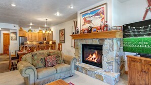 A cozy living room with a stone fireplace, a large TV, and a dining area with wooden cabinets and a chandelier. - Town Pointe C302 3 Bedroom Condo by RedAwning (Park City)