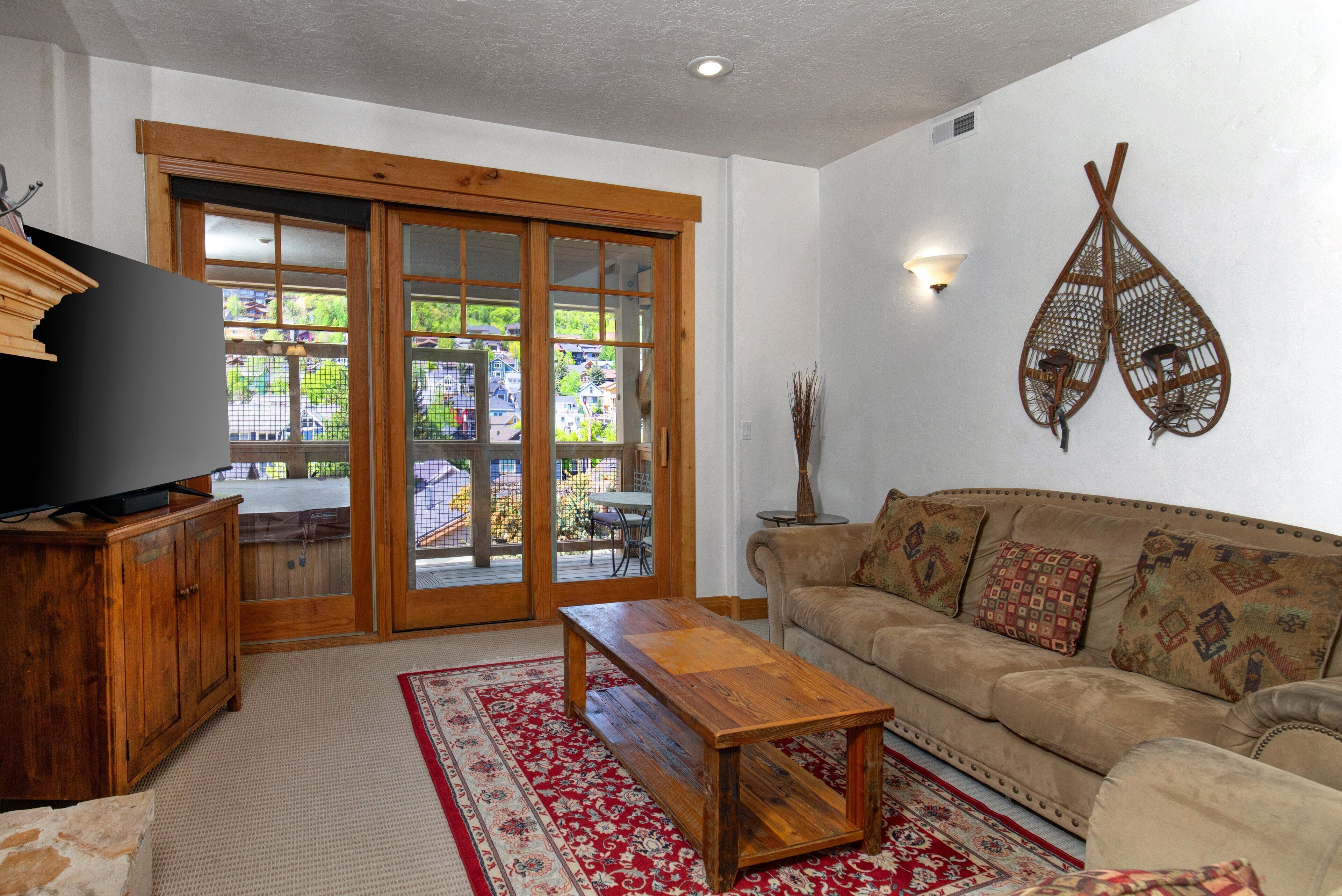 A living room with a wooden coffee table, a beige sofa with patterned cushions, a television on a wooden cabinet, and a large sliding glass door leading to an outdoor patio.