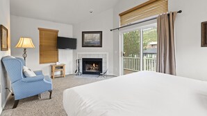 A hotel room with a large bed, a blue armchair, a fireplace, a television, and a balcony with a view of the outside.