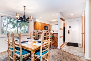 A dining area with a wooden table set for six, surrounded by matching chairs with teal cushions. A chandelier hangs above, and the room has wooden cabinets and a view through a large window. - Snow Flower 150 2 Bedroom Condo by RedAwning (Park City)