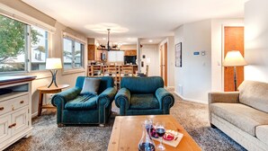 A spacious living room with two teal armchairs, a wooden coffee table with wine glasses, and a beige sofa. A kitchen with wooden cabinets and a dining area is visible in the background. - Snow Flower 150 2 Bedroom Condo by RedAwning (Park City)