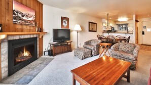 A living room with a fireplace, a television, patterned sofas, a wooden coffee table, and a dining area with chairs and a table. - Snow Flower 115 2 Bedroom Condo by RedAwning (Park City)