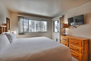A bedroom with a large bed, a wooden dresser, a television mounted on the wall, and a window with a view of trees. - Snow Flower 115 2 Bedroom Condo by RedAwning (Park City)