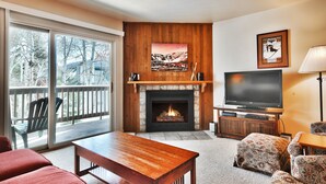 A living room with a fireplace, a television, and a balcony with a view of trees. - Snow Flower 115 2 Bedroom Condo by RedAwning (Park City)