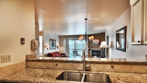 A kitchen with granite countertops, stainless steel sink, and a dishwasher. There is a dining area with a red sofa and a fireplace in the background. - Snow Flower 115 2 Bedroom Condo by RedAwning (Park City)