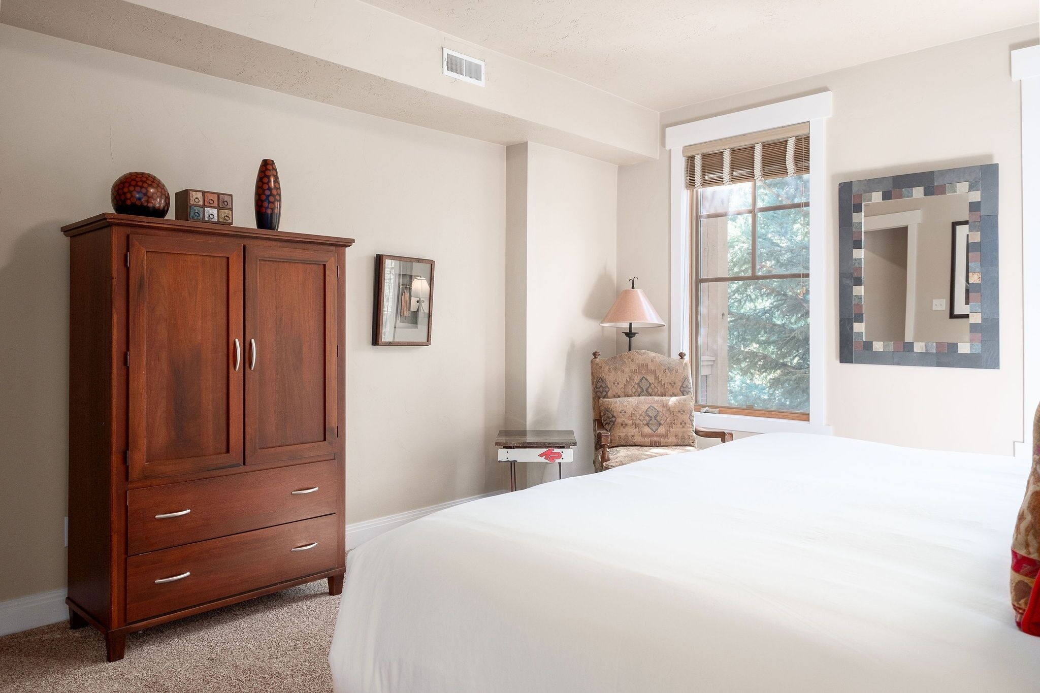 Quarto com uma cama grande, guarda-roupa de madeira, uma cadeira, mesa de cabeceira e janela com vista para árvores.