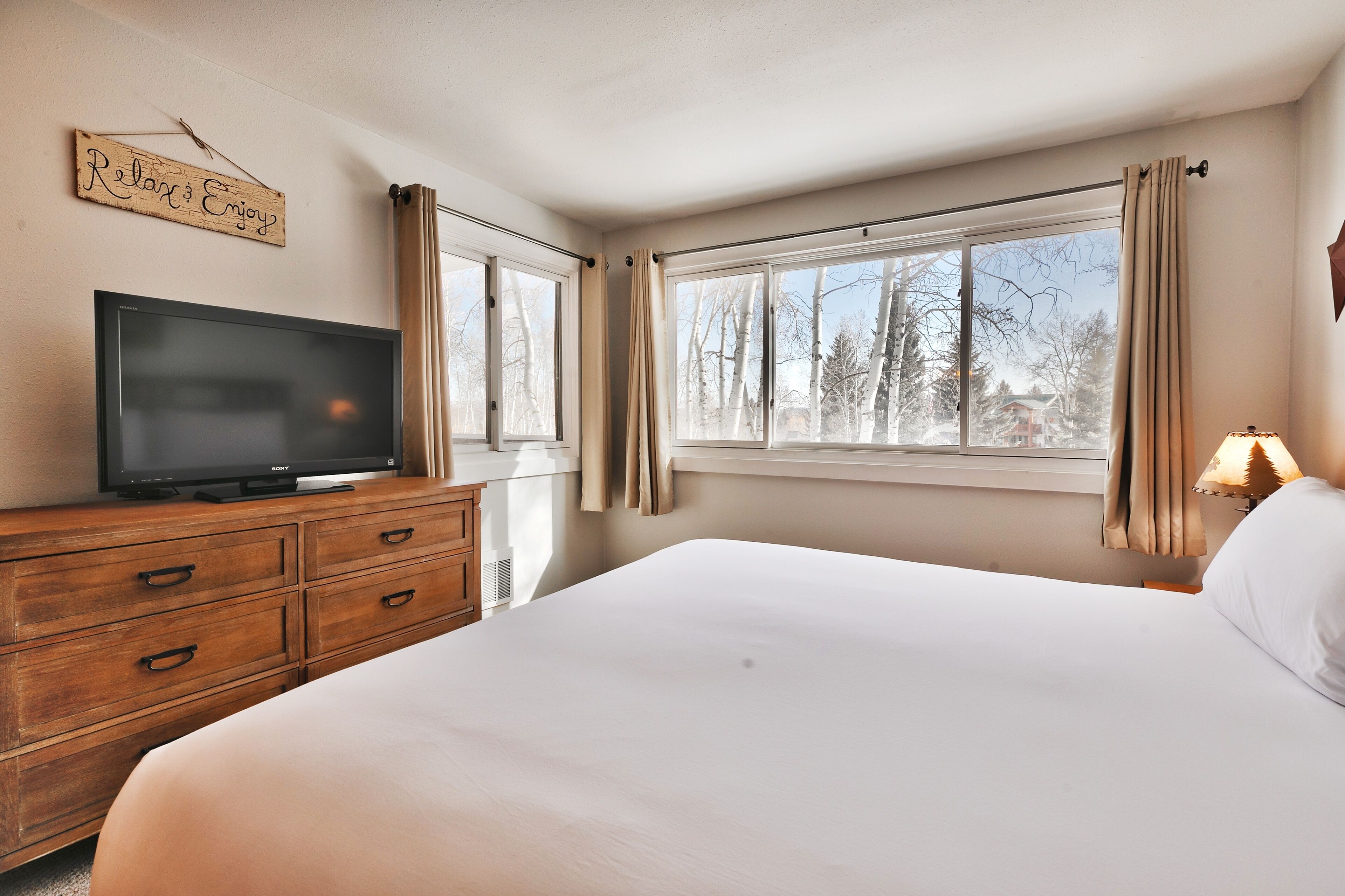 A bedroom with a bed, a TV on a wooden dresser, and a window with curtains.