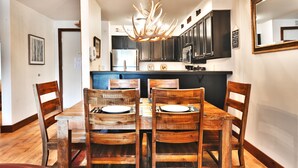 A dining area with a wooden table and chairs, a chandelier, and dark cabinetry in the background. - Snow Flower 104 2 Bedroom Condo by RedAwning (Park City)
