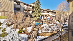 A multi-story building with a covered outdoor area, a staircase leading to a lower level, and snow on the ground. - Snow Flower 104 2 Bedroom Condo by RedAwning (Park City)