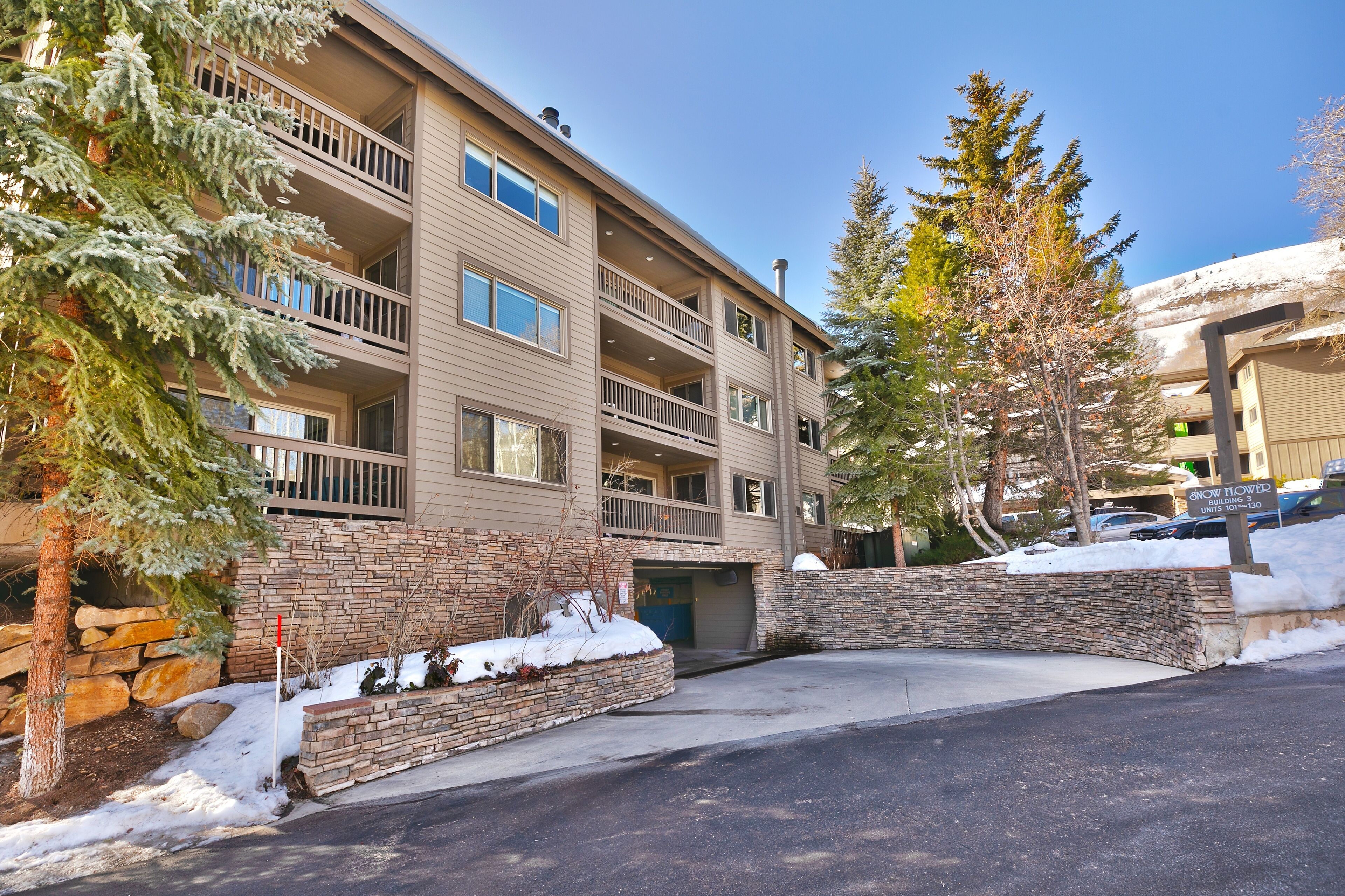 A multi-story building with balconies, a stone facade, and a parking area with snow.