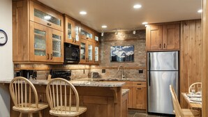 A kitchen with wooden cabinets, a stainless steel refrigerator, a microwave, and a stone backsplash. There are two wooden chairs and a dining table. - Snow Flower 042 1 Bedroom Condo by RedAwning (Park City)