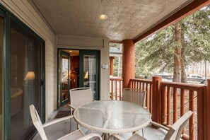 A covered outdoor seating area with a glass table and chairs, a wooden railing, and a view of trees and a parked car. - Snow Flower 042 1 Bedroom Condo by RedAwning (Park City)