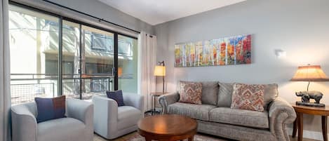 A modern living room with a large window, a sofa, two armchairs, a round wooden coffee table, and a painting on the wall.