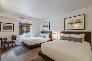 A hotel room with two beds, a desk, and a chair. There are framed pictures on the walls and a window with a view of trees.