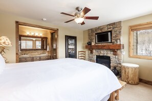 A bedroom with a large bed, a stone fireplace, a ceiling fan, and a television mounted above the fireplace. - Snow Flower 026 2 Bedroom Condo by RedAwning (Park City)