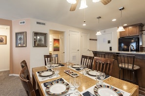Uma sala de jantar com mesa de madeira posta para seis pessoas, rodeada por cadeiras. Há uma cozinha ao fundo, com frigorífico e armários visíveis.
