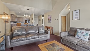 A spacious living area with a leather sofa, a wooden coffee table, and a kitchen in the background. - Snow Flower 127 2 Bedroom Condo by RedAwning (Park City)