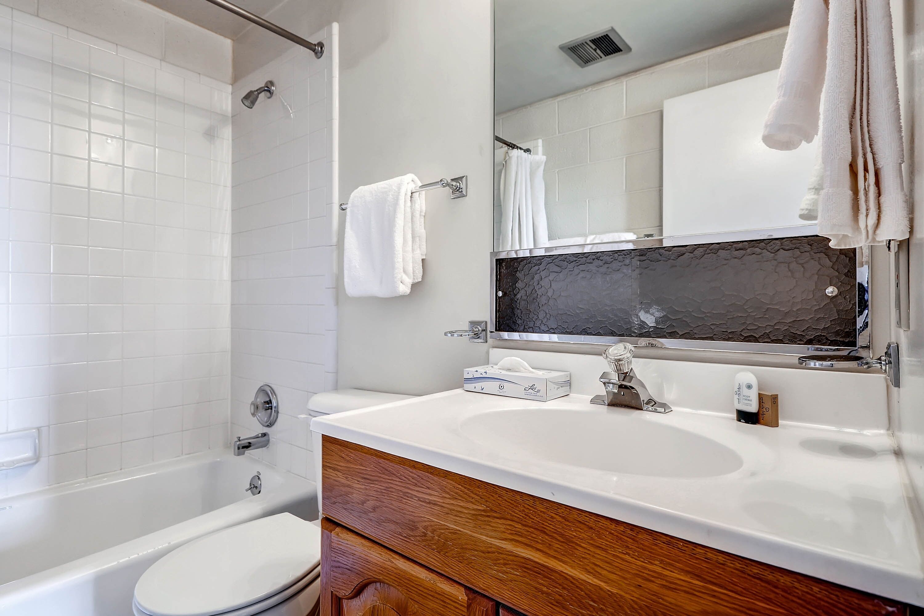 Un baño con una bañera blanca, un lavamanos blanco, un tocador de madera y un espejo con un salpicadero negro.