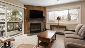 A living room with a fireplace, a television, a sofa, a coffee table, and a balcony with outdoor furniture. - Snow Flower 113 2 Bedroom Condo by RedAwning (Park City)