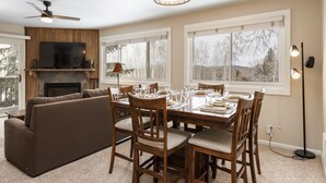 A spacious living room with a fireplace, a dining area with a table set for four, and a large TV mounted on the wall. - Snow Flower 113 2 Bedroom Condo by RedAwning (Park City)