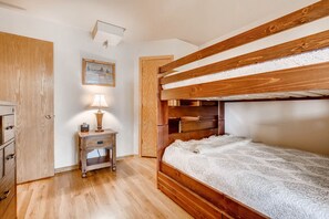 A bunk bed with a wooden frame and a white textured mattress. There is a wooden nightstand with a lamp and a small table. A wooden wardrobe is visible on the left.