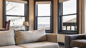 Una sala de estar con un sofá, una silla y una mesa. Hay ventanas amplias con vistas al exterior.