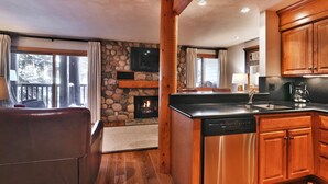 A cozy kitchen with wooden cabinets, a stone fireplace, and a large window overlooking a deck. - Snow Flower 003 2 Bedroom Condo by RedAwning (Park City)