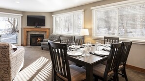 A dining area with a fireplace, a television mounted above it, and large windows with blinds.