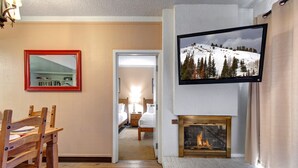 Habitación de hotel con una mesa de comedor de madera, un televisor sobre la chimenea y un pasillo que conduce a un dormitorio.