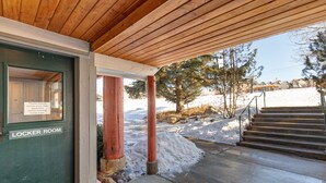 Une entrée couverte en bois mène à un bâtiment sur lequel est apposée une enseigne indiquant « VESTIAIRES ». On peut apercevoir un paysage enneigé et un escalier menant à un autre bâtiment.
