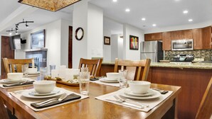 A dining area with a wooden table set for four, surrounded by wooden chairs. A kitchen with wooden cabinets and stainless steel appliances is visible in the background. - Park Station 113 2 Bedroom Condo by RedAwning (Park City)