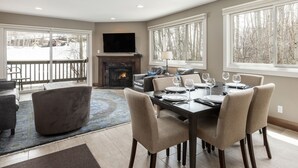 A dining area with a fireplace, a television, and large windows with a view of trees. - Snow Flower 120 2 Bedroom Condo by RedAwning (Park City)