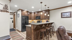 A modern kitchen with wooden cabinets, a black refrigerator, a microwave, and a bar area with stools. - Snow Flower 071 3 Bedroom Condo by RedAwning (Park City)