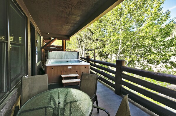 Una terraza de madera con jacuzzi, mesa de vidrio y sillas, con vistas a un área boscosa.