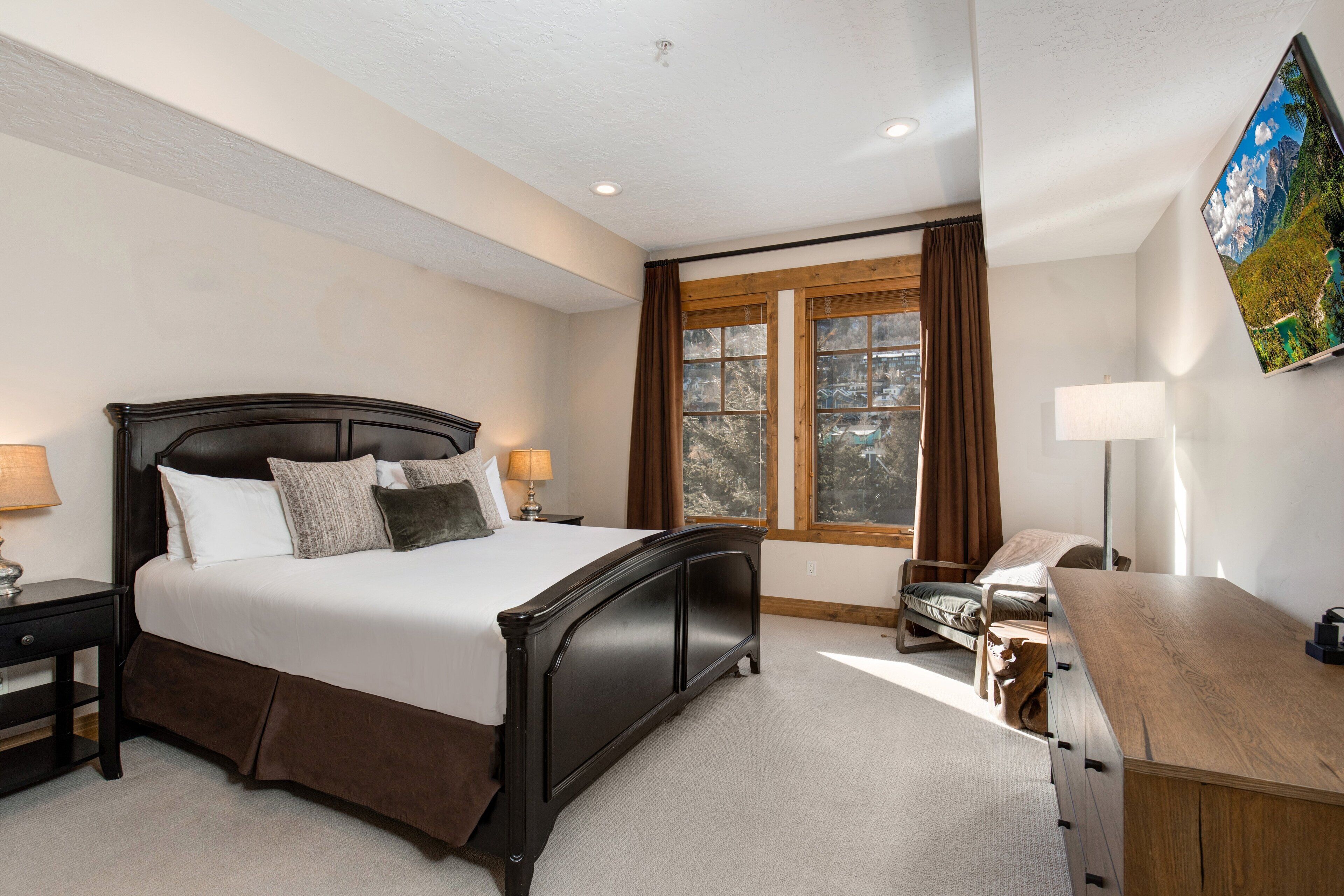 A bedroom with a large bed, a wooden desk, a nightstand, and a window with curtains.