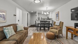 A modern living room with a brown sofa, a wooden coffee table, and a beige armchair. A dining area with a kitchen in the background is visible. - Snow Flower 134 2 Bedroom Condo by RedAwning (Park City)